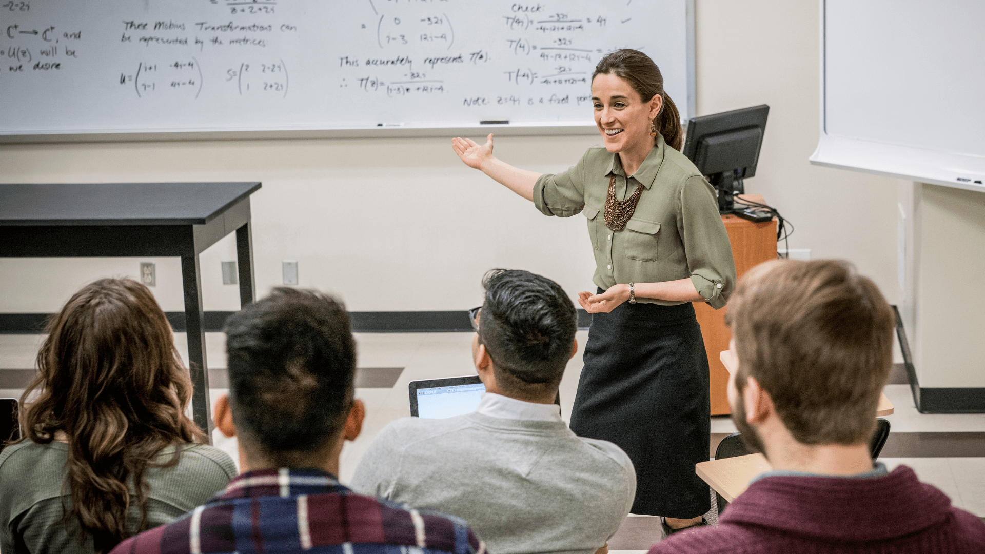 A female teacher talking to a class and pointing at a whiteboard