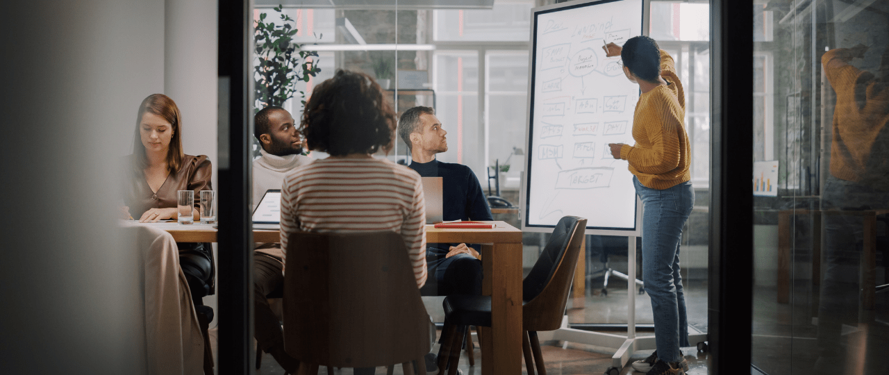 A group of people sat in a room working with one person stood up pointing at a whiteboard