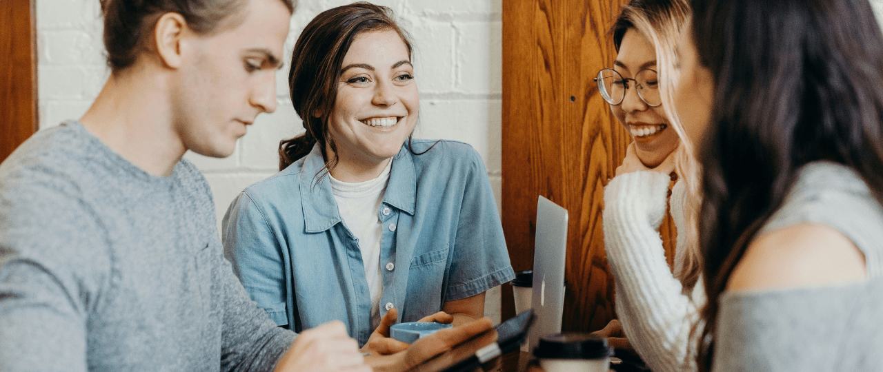 Four young people smiling sat at a table in conversation and looking at a tablet device