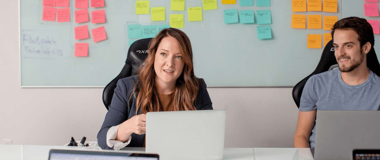 A man and woman sat at a desk working on laptops with a wall of post it notes behind them