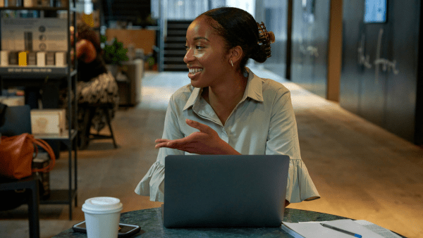 A young woman sat at a table on her computer talking to someone next to her