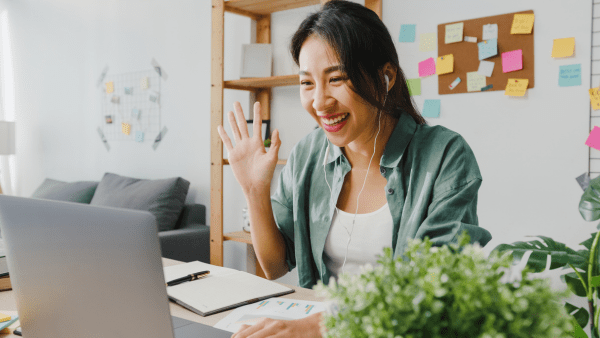 A young woman smiling and waving as she takes a virtual meeting on her computer