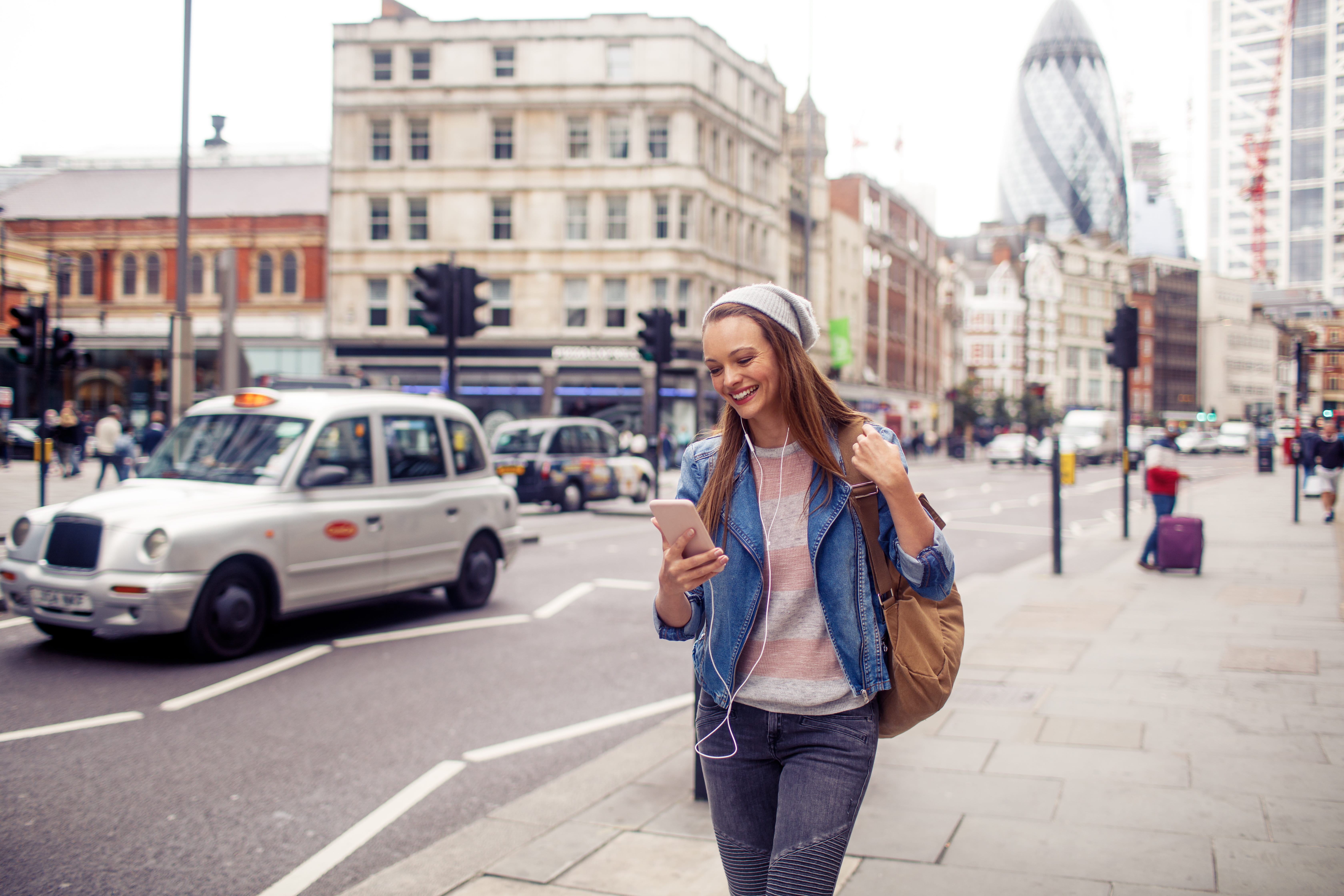 woman walking down street with earbuds in and looking at her cell phone