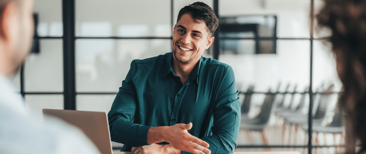 A man sat at a table smiling and presenting across from two people 