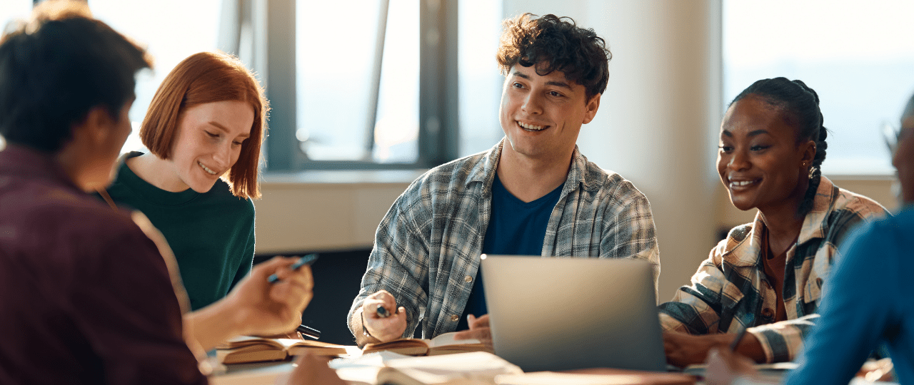 A group of students sat around a table studying and smiling while looking at a laptop