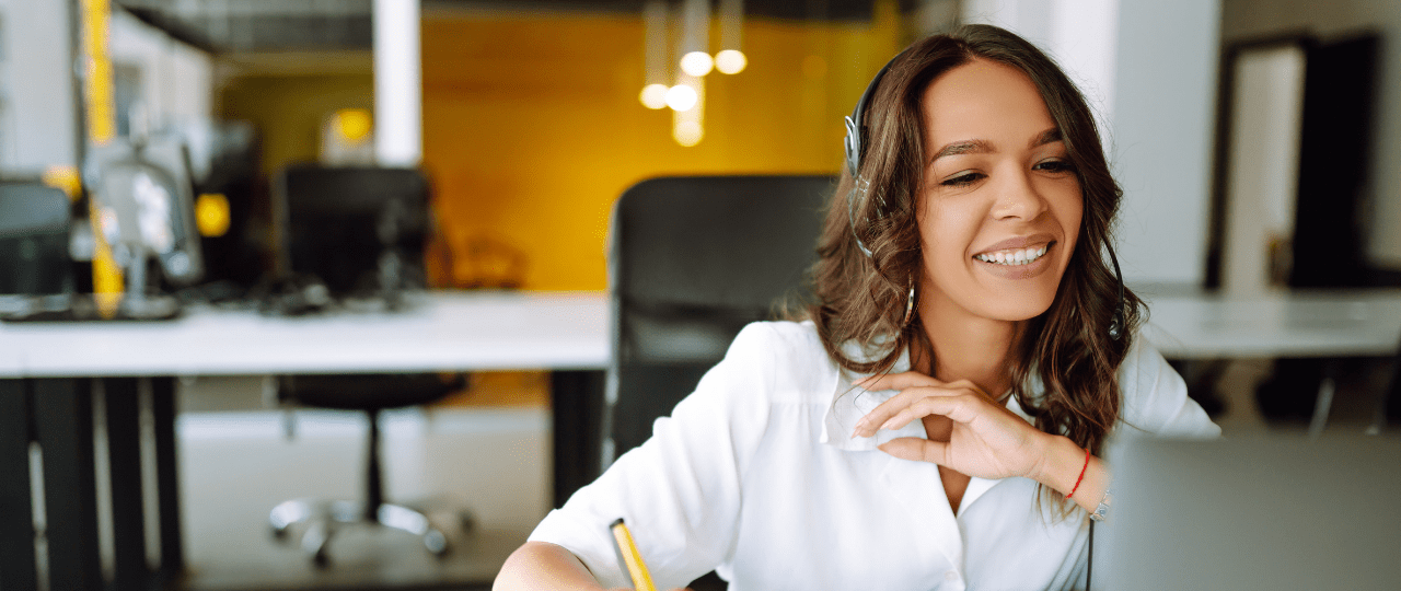 Woman sat a a desk smiling while writing with a pen and looking at a computer