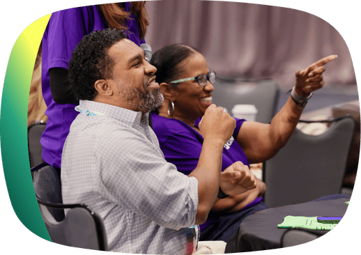 Man sits at a table with a woman in a purple t-shirt and glasses pointing