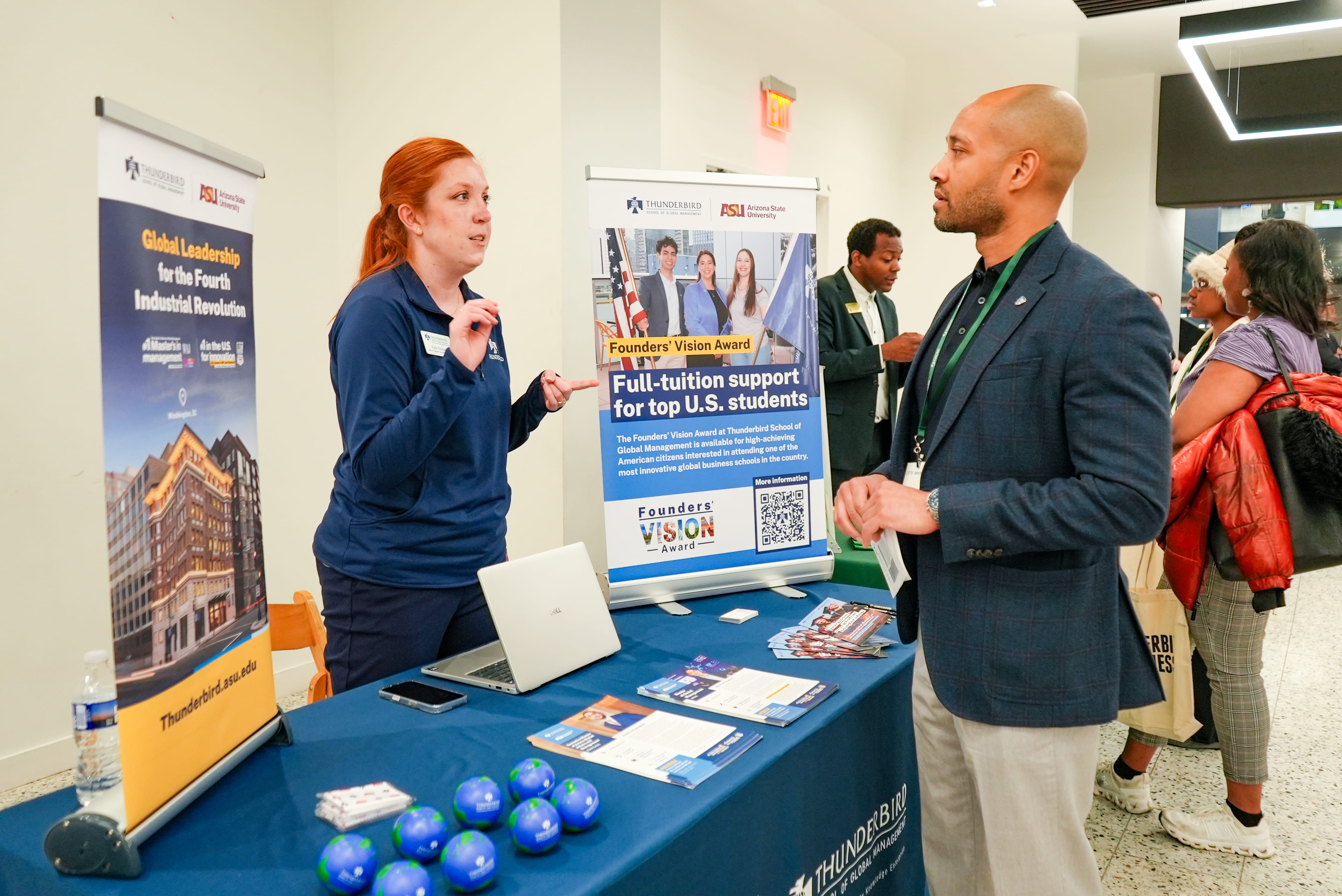 Women talks to a man in a blazer from behind a table with a blue tablecloth and pop-up signs