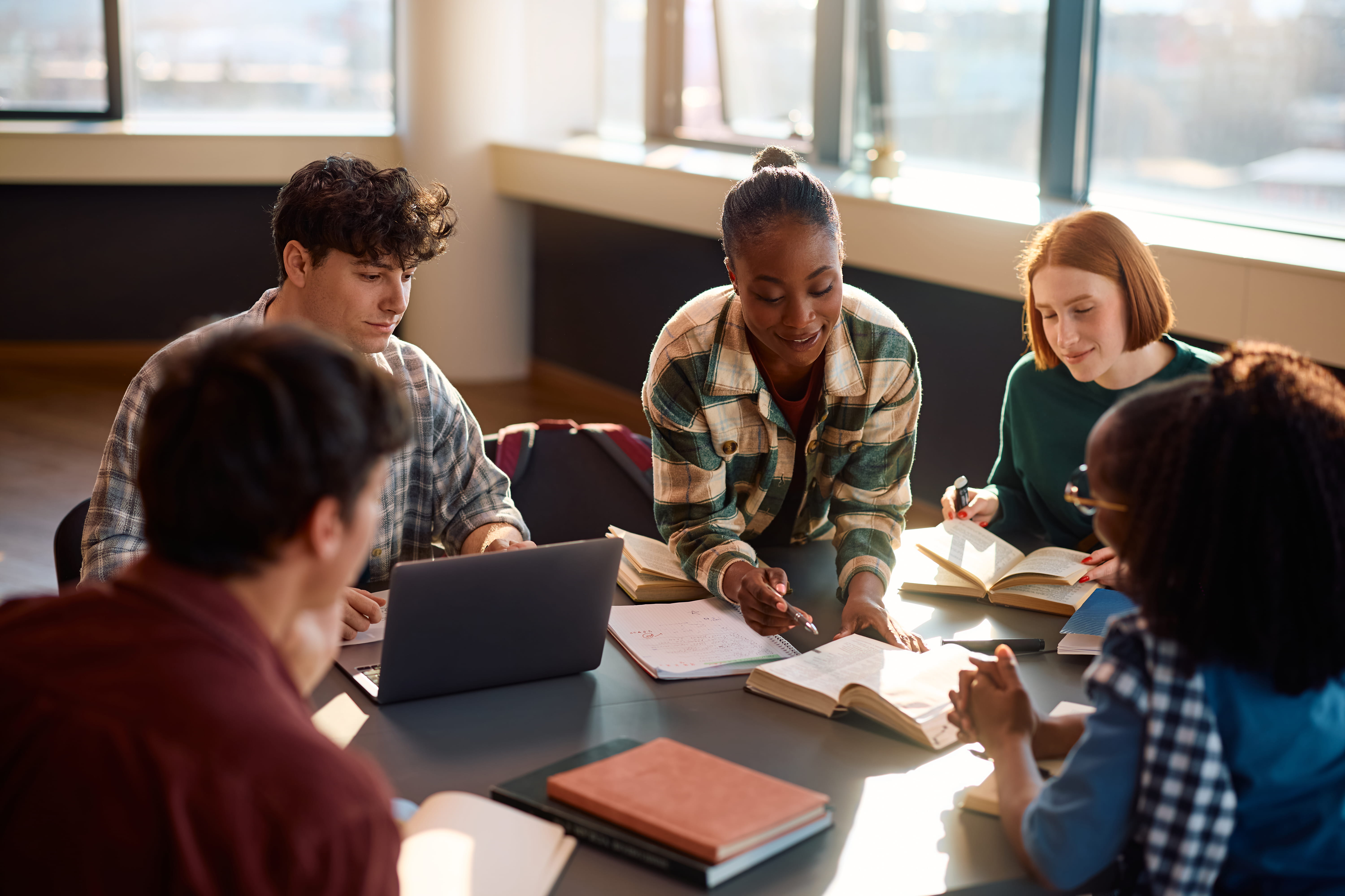 Woman points to book while sitting at a table with four other classmates