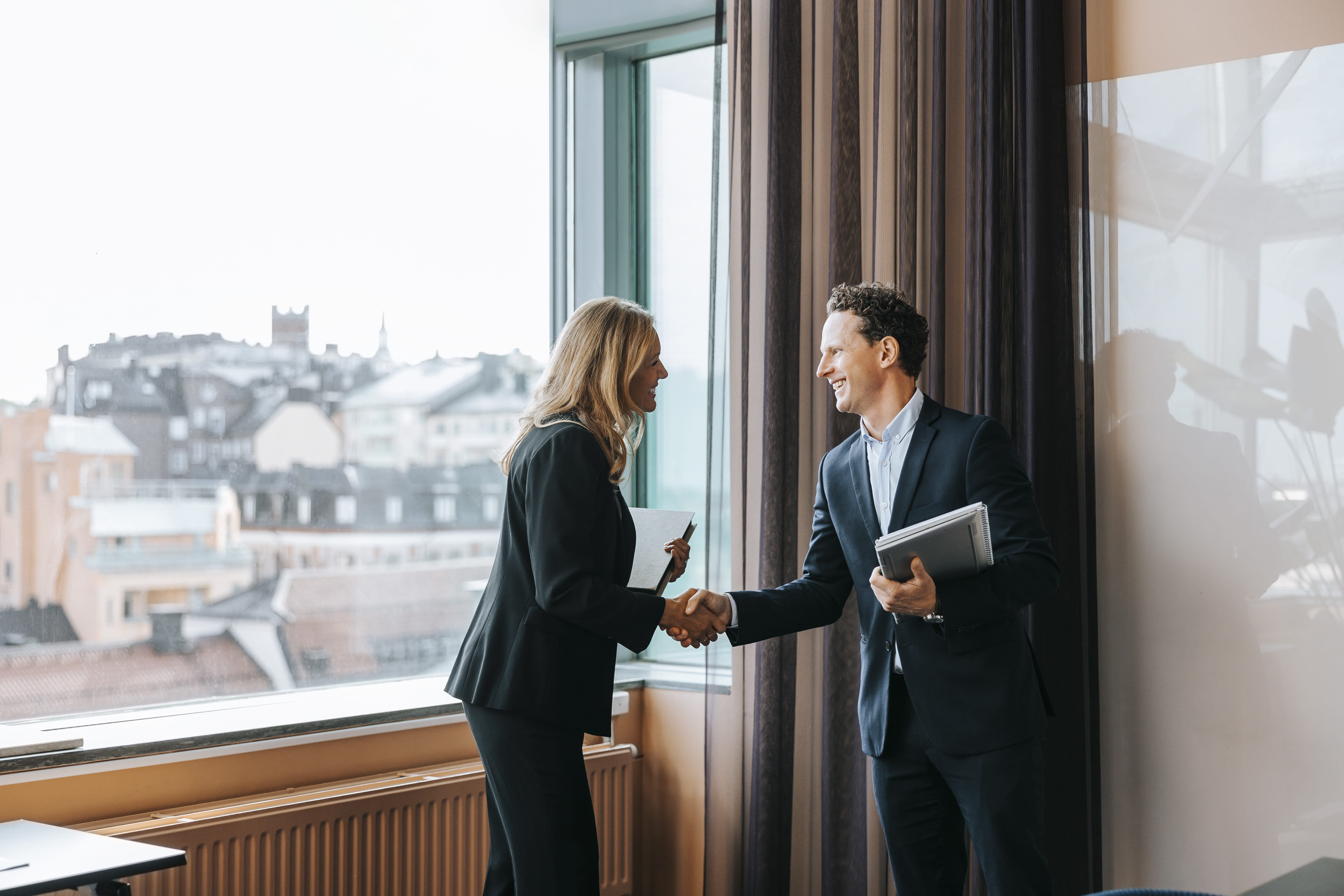 Woman and man wearing business attire shaking hands in front of a window