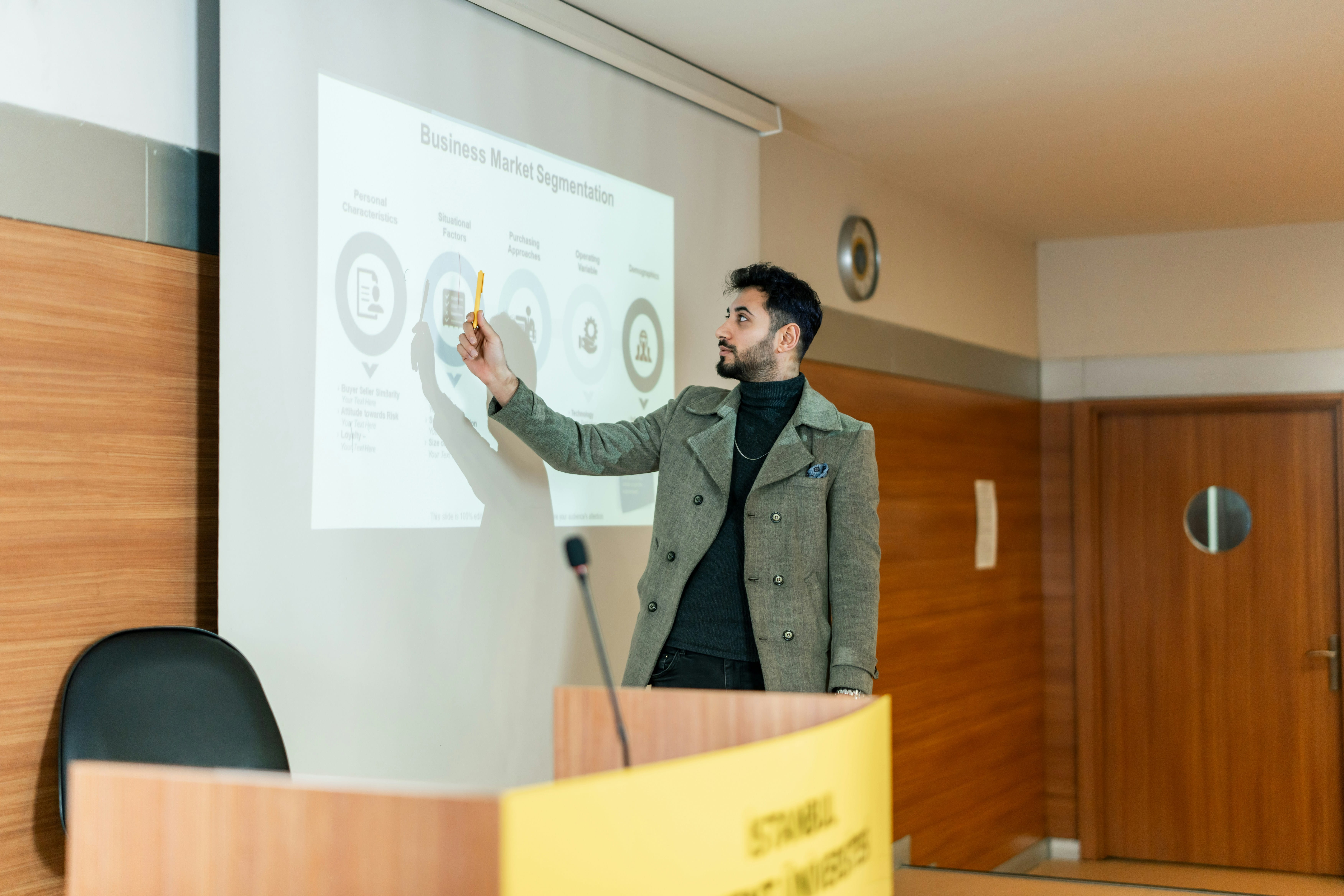 Man teaching at the front of a classroom pointing to a graph on a projector screen